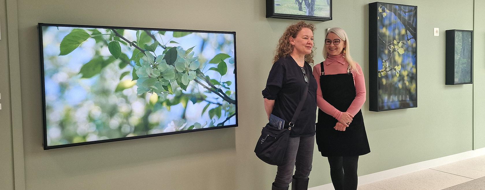 Kaksi naista seisoo suuressa tilassa. Taustalla vihreä seinä, jossa valokuvia puista.