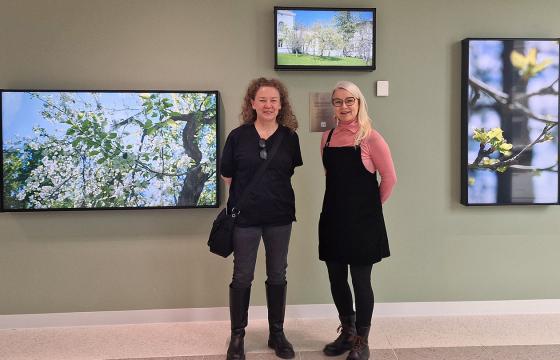 Kaksi naista seisoo suuressa tilassa. Taustalla vihreä seinä, jossa valokuvia puista.