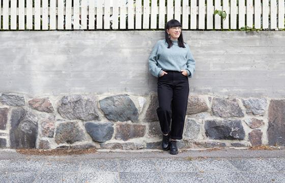 Tummahiuksinen nainen mustissa housuissa ja sinisessä paidassa nojaa muuriin.