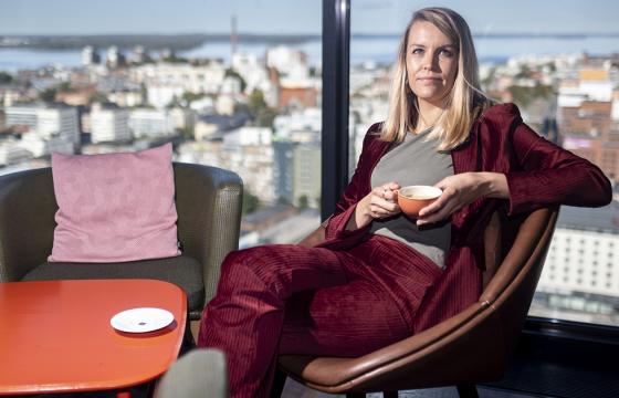 Nainen punaisessa samettijakussa istuu kahvikupin kanssa nojatuolissa. Taustalla ikkunasta näkyy kaupungin silhuetti.