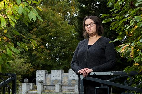 Mustapaitainen ja tummatukkainen nainen seisoo vihreän hautakivien edustalla ja nojaa aitaan.