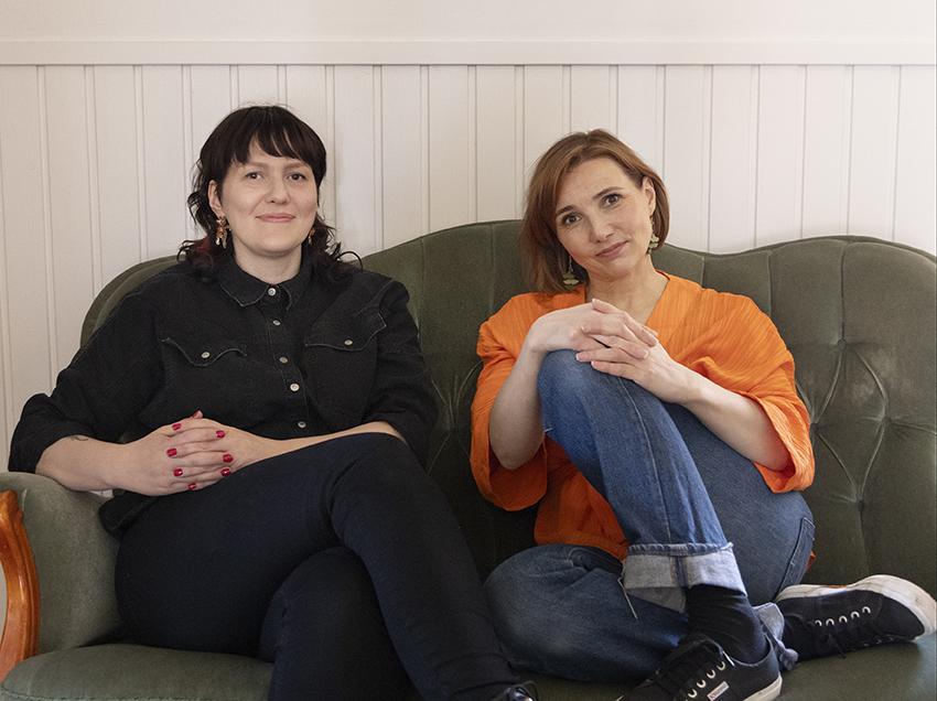 Kaksi naista istuu vierekkäin vihreällä sohvalla katsoen kameraan