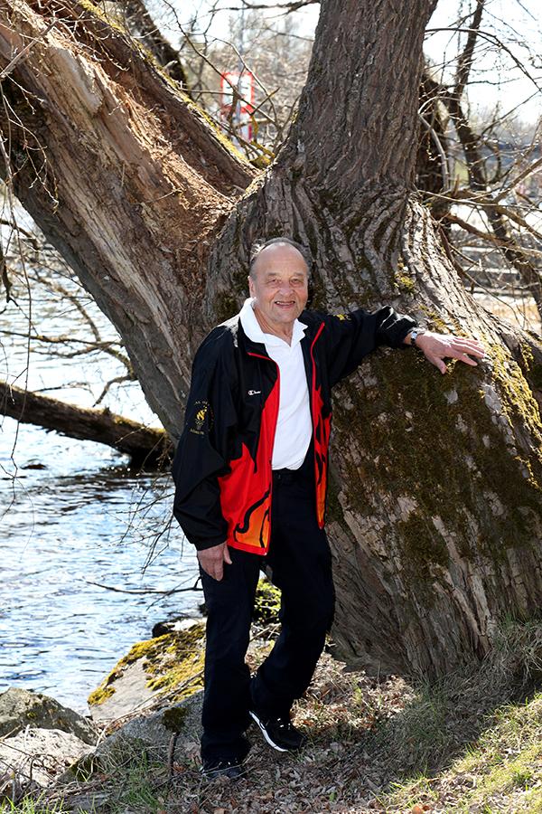 Mies seisoo joen rannassa ja nojaa vanhaan puuhun.