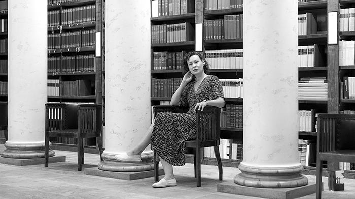 Mustavalkoinen kuva, nainen istuu tuolilla kirjahyllyn edessä.