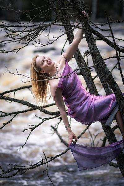 Nainen violetissa juhlamekossa roikkuu puun oksasta kuohuvan kosken äärellä.