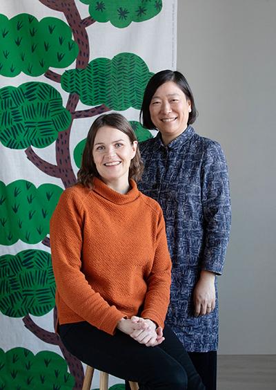 Kaksi naista seisoo vierekkäin, taustalla kangas, jossa vihreä puu