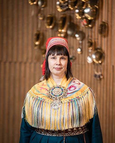 Nainen saamelaisessa kansallispuvussa. Taustalla kullanvärisiä taideteoksia