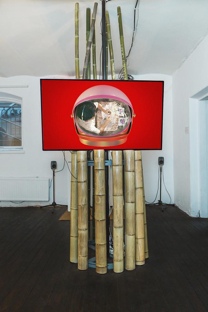 Monika Czyżyk, Geographies of a lonely Pole (I want to build a rocket), 2020, 4K UHD video, 82 min 48 s, color, stereo sound, 55” screen, auto pole mounting, bamboo poles with carved drawings by Torsten Zenas Burns mounted on steel hoop rack, 286 x 60 x 70 cm. Myymälä2 gallery. Photo: Laura Mainiemi.