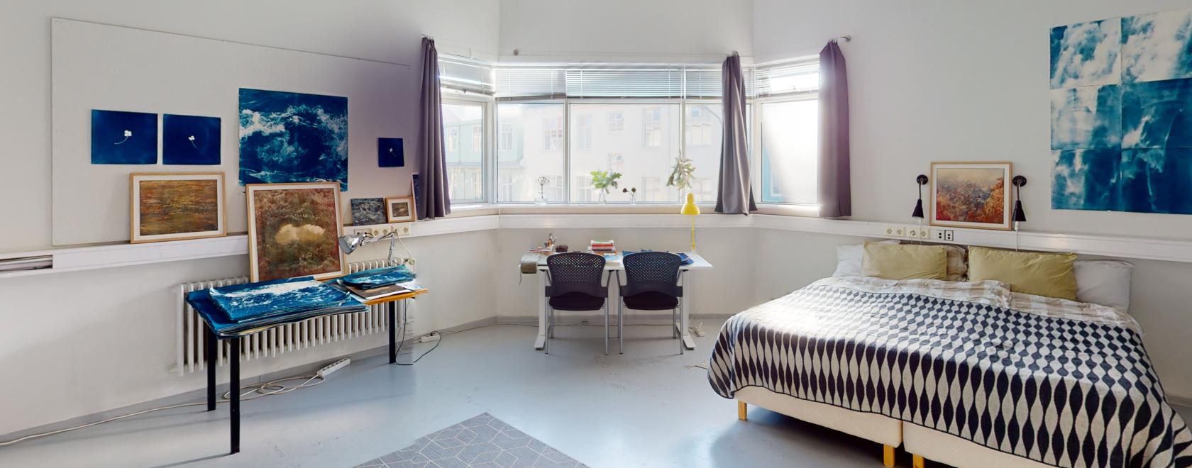Kuva vaaleasta huoneesta, jossa on leveä ikkuna, sänky ja kaksi pöytää.