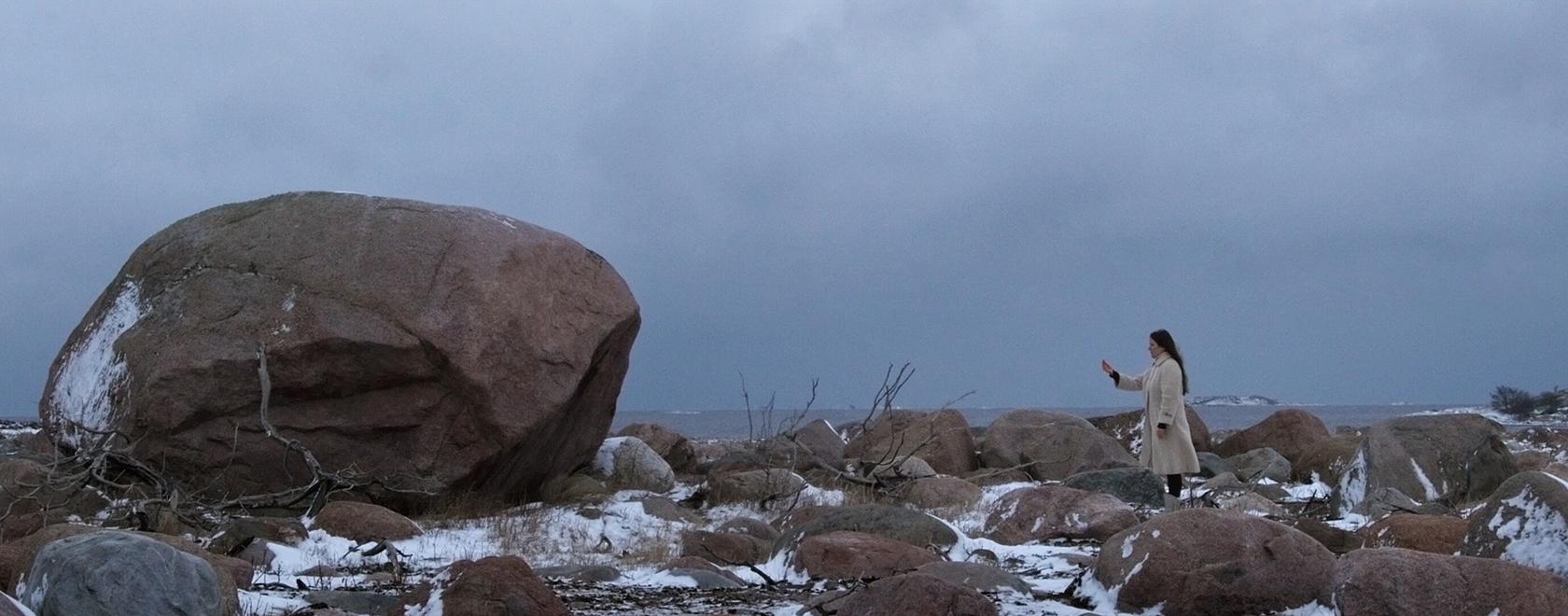 Valokuva talvisesta maisemasta, vasemmalla suuri kivi, oikealla pienessä koossa ihminen