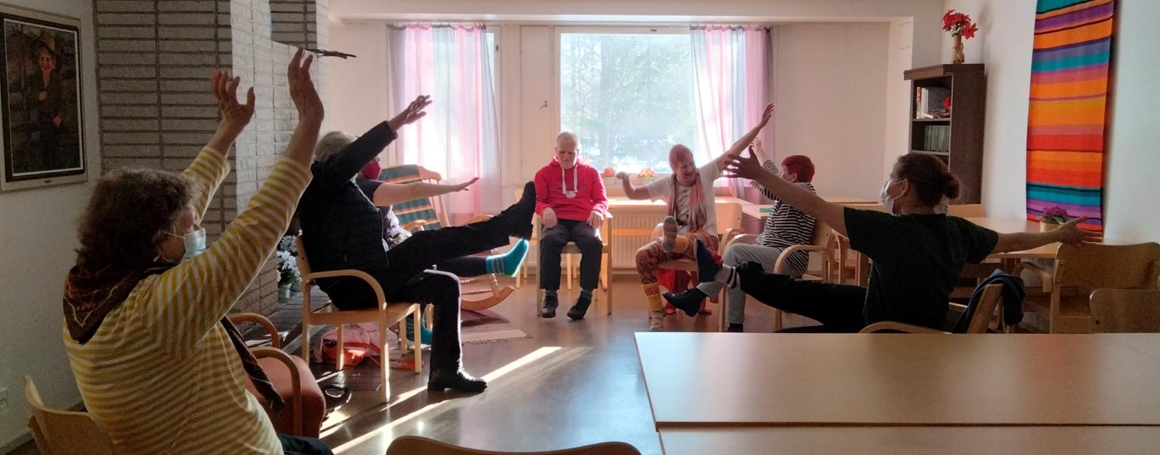 Vanhoja ihmisiä istumassa tuoleilla ympäri huonetta. Ihmiset tanssivat istualtaan.