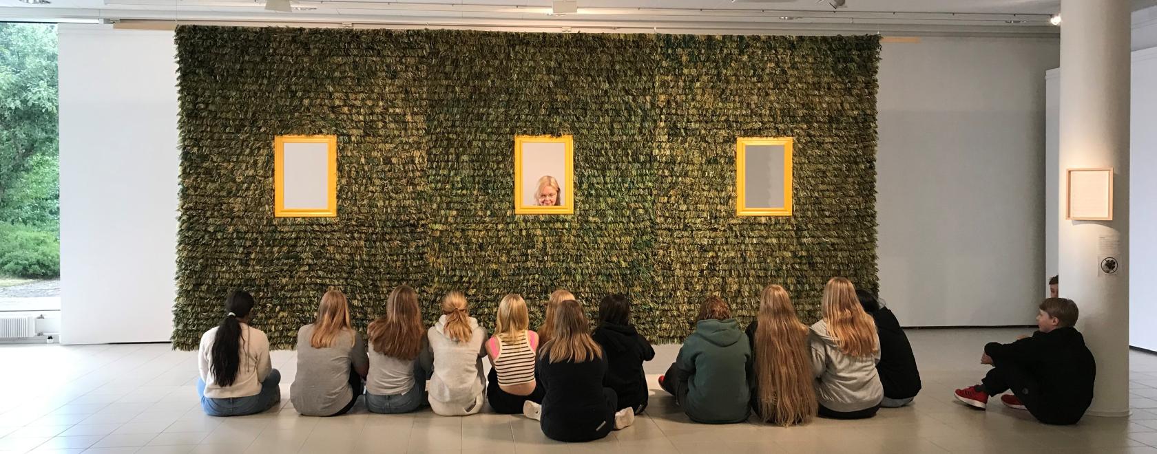 Nuoria istuu lattialla rivissä selät kameraan. He katsovat seinällä olevaa vihreää taideteosta.