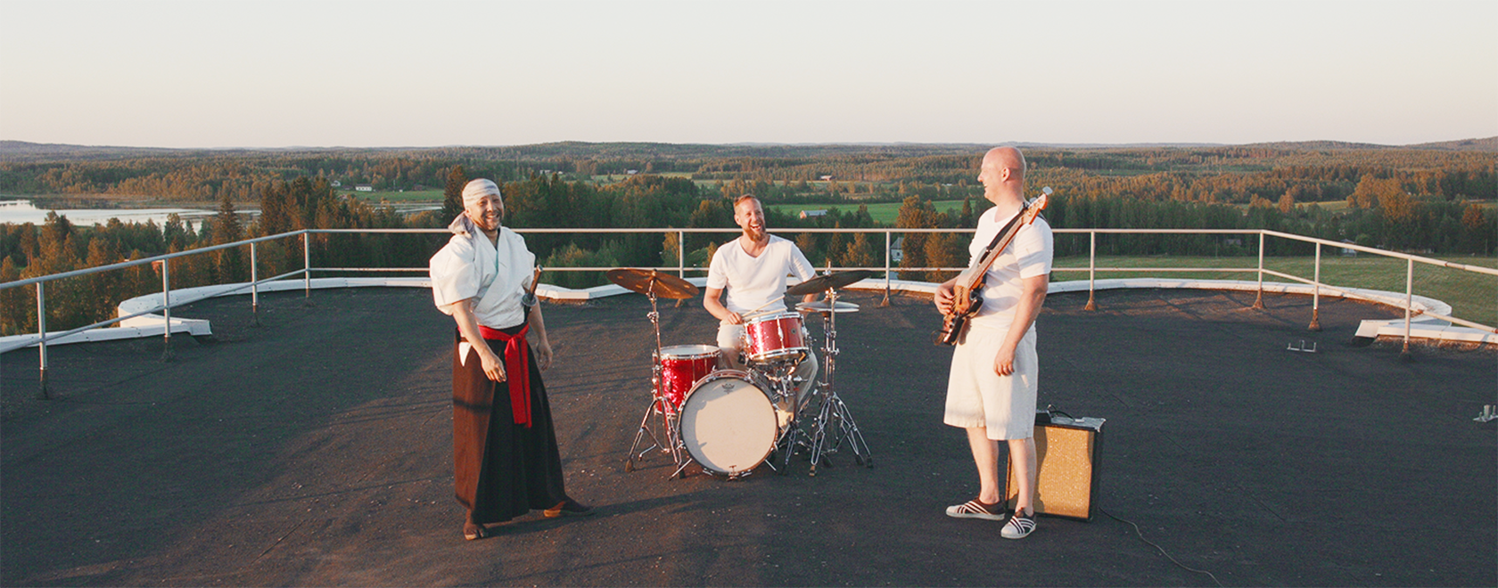 Kolme miestä seisoo katolla ja soittavat rumpuja ja kitaraa. Taustalla kesäinen maisema.
