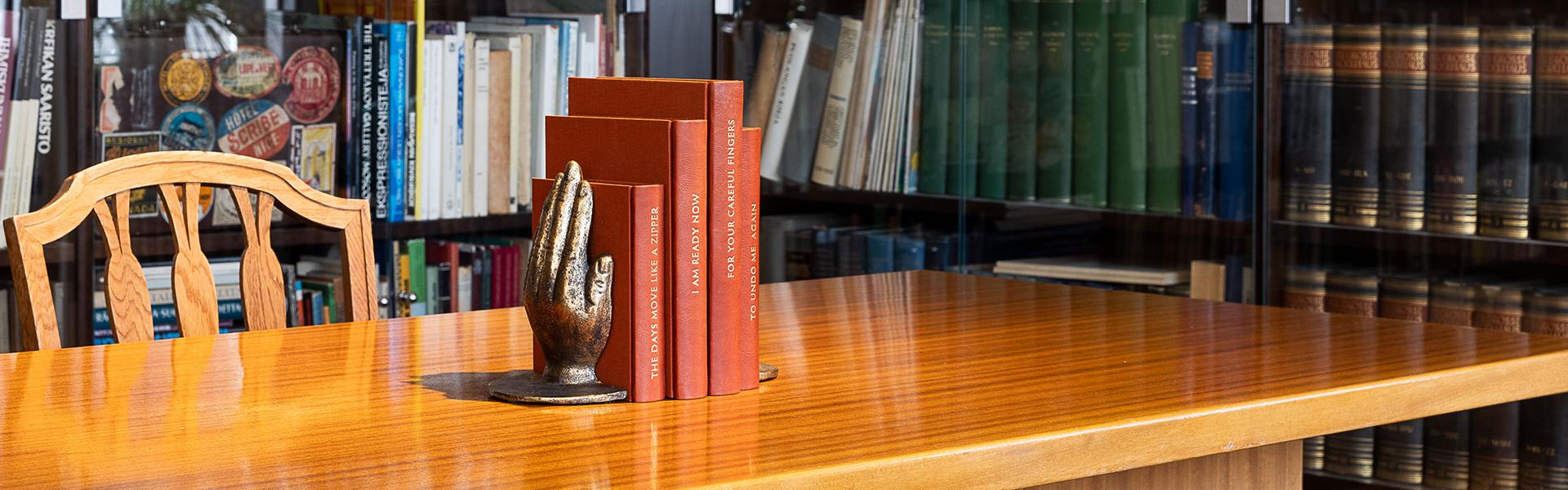 Ruskea pöytä, jossa kädenmuotoinen kirjanpidike ja punaisia kirjoja. Taustalla kirjahylly.