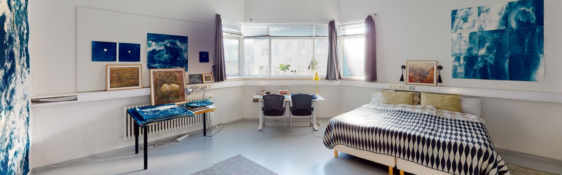 Kuva vaaleasta huoneesta, jossa on leveä ikkuna, sänky ja kaksi pöytää.