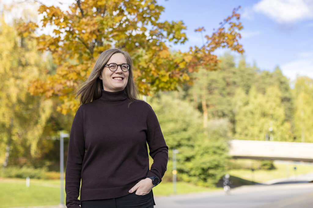 Tummissa vaatteissa oleva nainen seisoo ulkona syksyisessä maisemassa