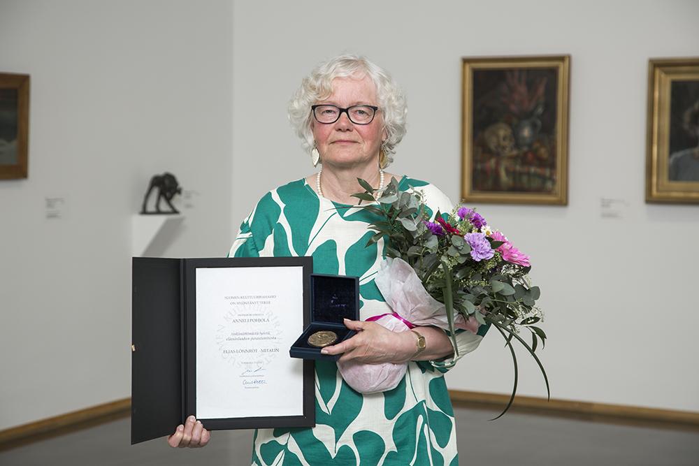 Nainen seisoo taidegalleriassa kukkakimppu ja diplomi kädessään.