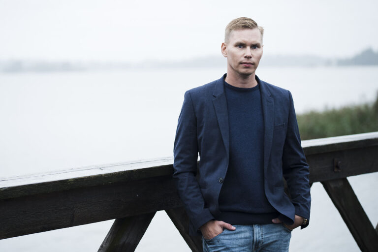 Mies seisoo puvuntakki päällä nojaten sillan kaiteeseen. Taustalla järvimaisema