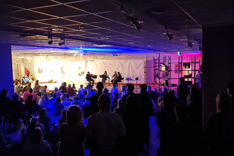 Hämärä kuva, lavalla soittajia ja edessä paljon yleisöä
