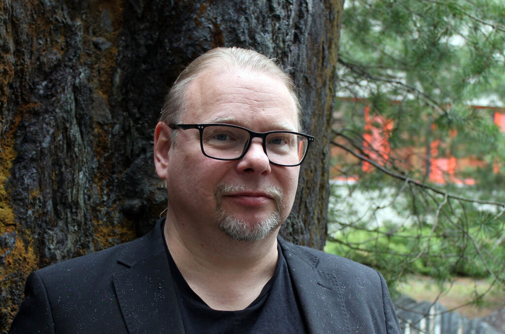 Silmälasipäinen mies seisoo puunrungon edessä