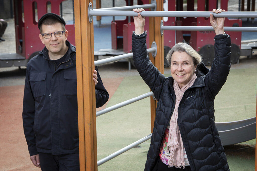 Mies ja nainen nojailemassa hymyillen lasten leikkikentän kiipeilytelineeseen.