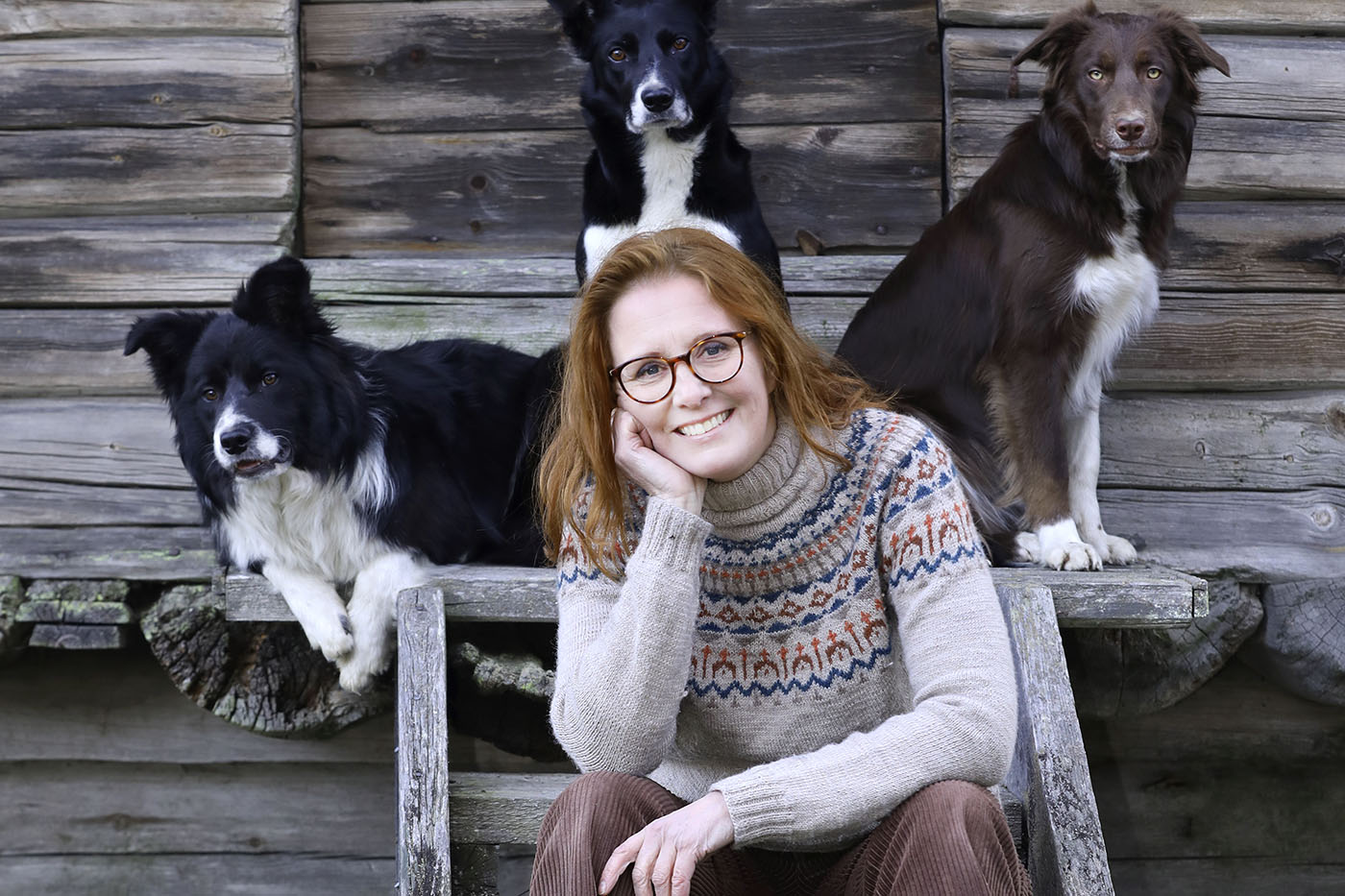 Nainen istuu vanhan hirsitalon rappusilla kolmen koiran kanssa.