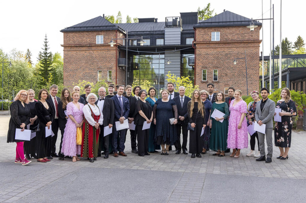 Suuri joukko ihmisiä juhlapuvuissaan ulkona. Taustalla punatiilinen rakennus, jossa on musta harjakatto.