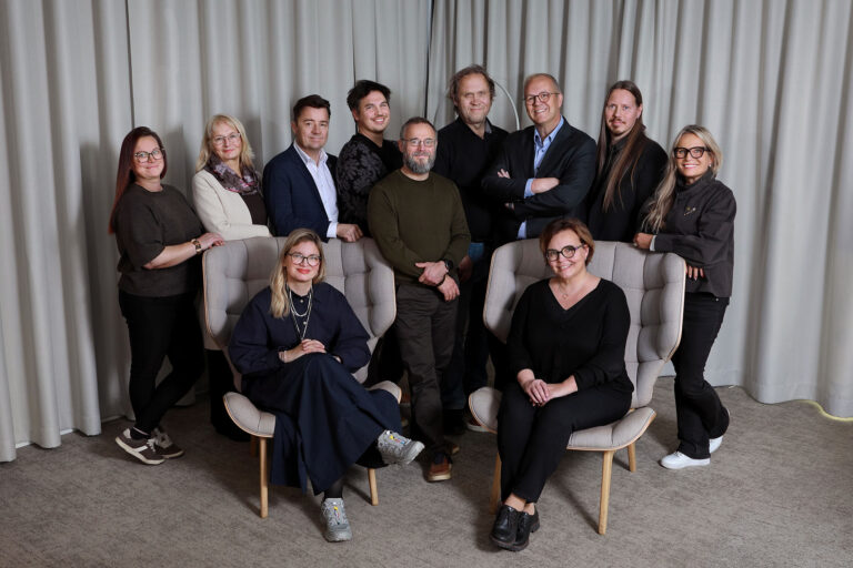 Iso joukko ihmisiä studiossa, taustalla harmaa kangas