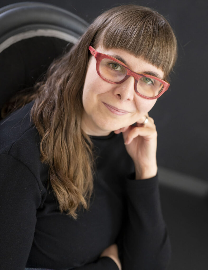 Nainen mustassa paidassa ja punaisissa silmälaseissa nojaa kämmeneensä ja katsoo hymyillen kameraan.
